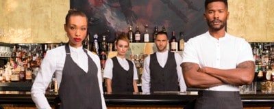 Das Bild zeigt vier Personen, die offenbar als Barpersonal gekleidet sind, vor dem Hintergrund einer gut bestückten Bar.