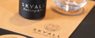 Das Bild zeigt einen Teil einer Menükarte und ein Wasserglas mit einem Logo, das sich auf den Namen "SKYFALL Beach Lounge & Bar" bezieht.